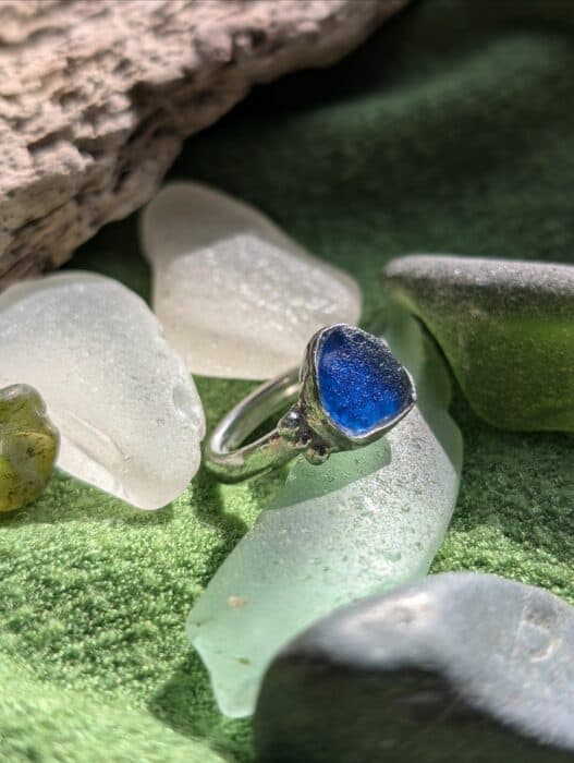 A handcrafted fine silver ring featuring a rare, vibrant cobalt blue sea glass stone. The ring is photographed outdoors on a mossy green surface surrounded by translucent sea glass pieces.