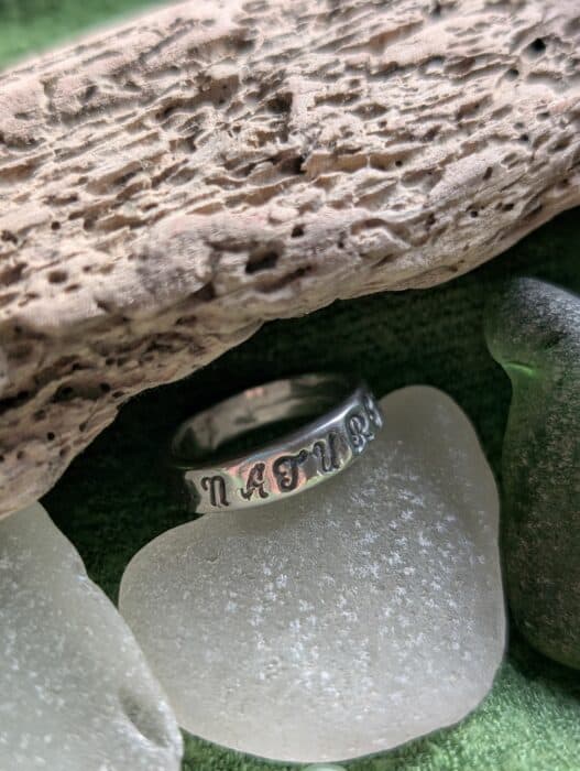 A wide, handcrafted fine silver ring with the word 'NATURE' hand-stamped into the polished metal, resting against a textured driftwood background