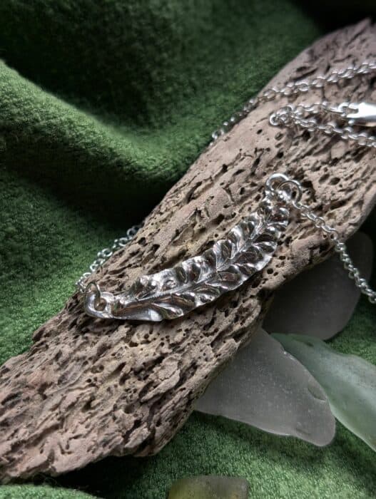 A detailed fine silver pendant in the shape of budding leaf, showing intricate botanical textures, resting on a piece of weathered driftwood with a silver chain.