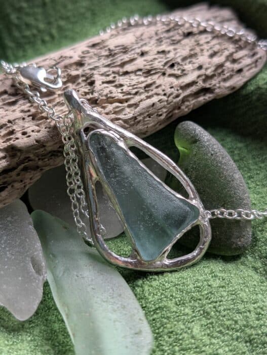A long, triangular piece of pale frosted sea glass with a natural horizontal line through the middle, encased in a bespoke, hand-forged fine silver frame on a silver chain.