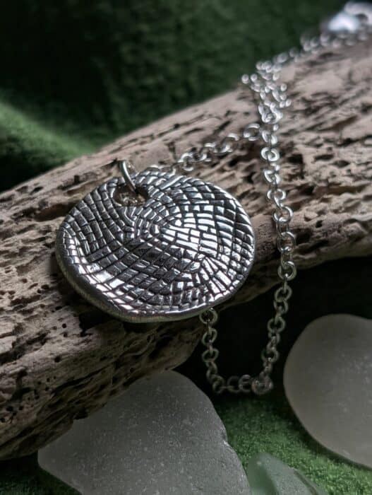 A circular fine silver pendant with a complex, woven spiral texture that catches the light, resting on a weathered piece of driftwood with a delicate silver chain.
