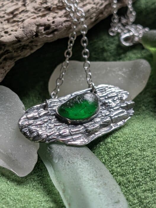 A handcrafted fine silver pendant with a deep, bark-like driftwood texture holding a vibrant, translucent green sea glass stone. The silver frame securely cradles the glass on a natural wood background.